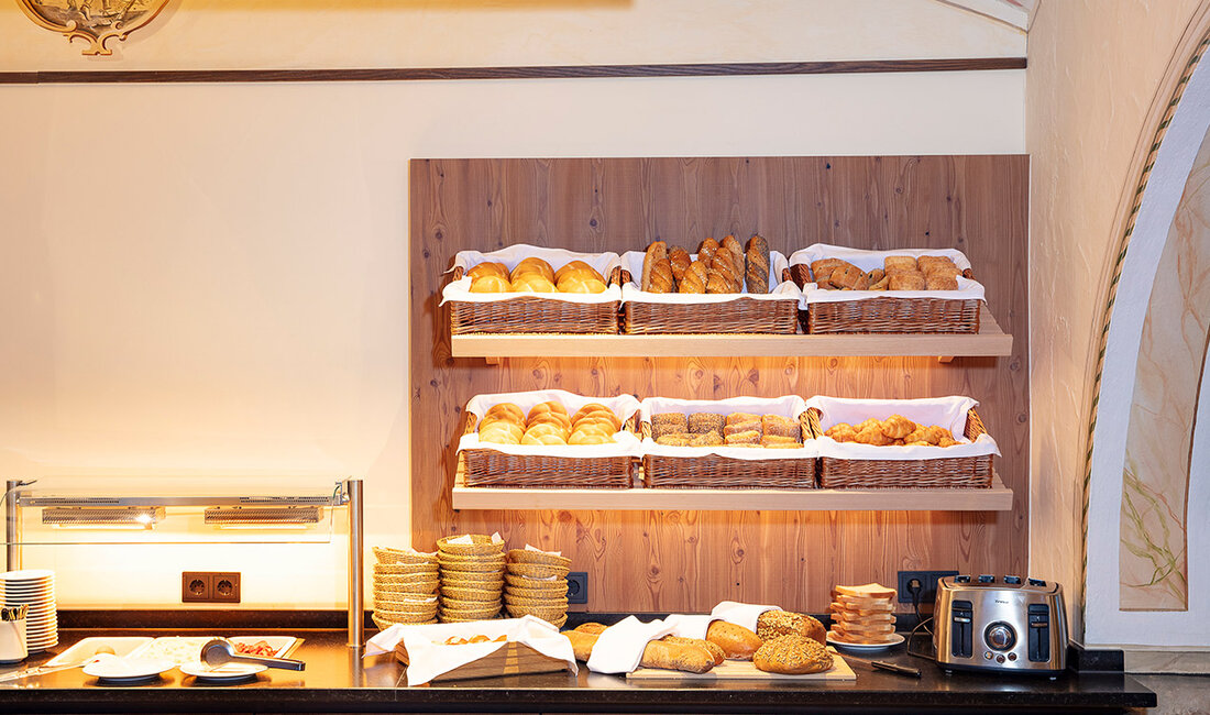 Ein Frühstücksbuffet mit Körben voller verschiedener Backwaren, Tellern, einem Toaster und einer Warmhalteplatte auf einer Holztheke, die an einer Wand mit Zierleisten steht.