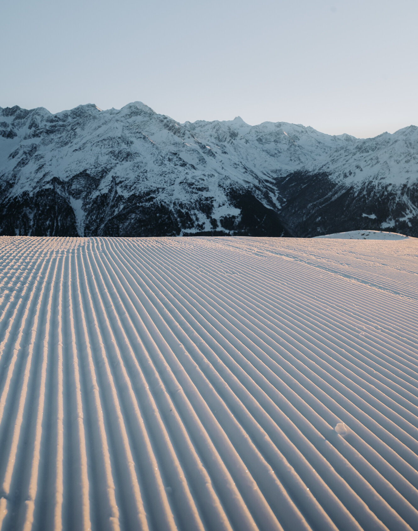 Rillenförmiger Schnee bedeckt ein weites Feld, das bei Sonnenaufgang oder Sonnenuntergang unter klarem Himmel zu den fernen, schneebedeckten Bergen hinführt.