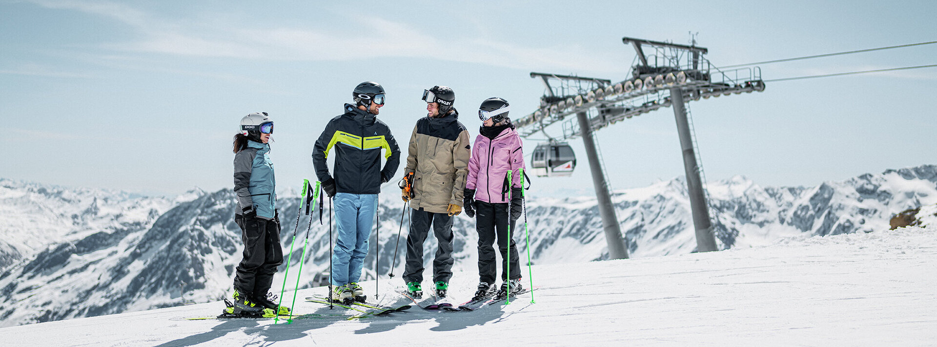 Vier Personen in Skikleidung stehen gemeinsam mit Skistöcken auf einem verschneiten Berg. Im Hintergrund sind unter einem strahlend blauen Himmel ein Skilift und schneebedeckte Gipfel zu sehen.