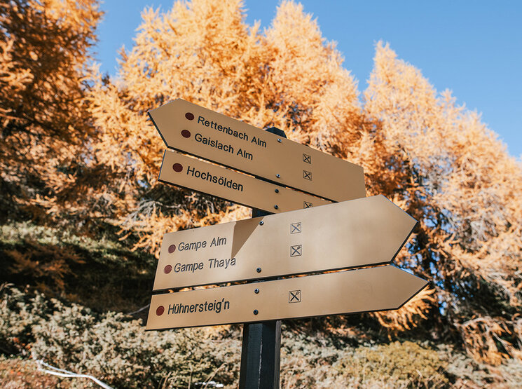 Gelbe Wanderwegschilder mit schwarzer und roter Beschriftung weisen in verschiedene Richtungen und heben sich vor dem Hintergrund herbstlicher Bäume mit orangefarbenen Blättern und einem strahlend blauen Himmel ab. Auf den Schildern sind Ziele wie Hochsölden, Rettenbach Alm und Hühnersteign angegeben.