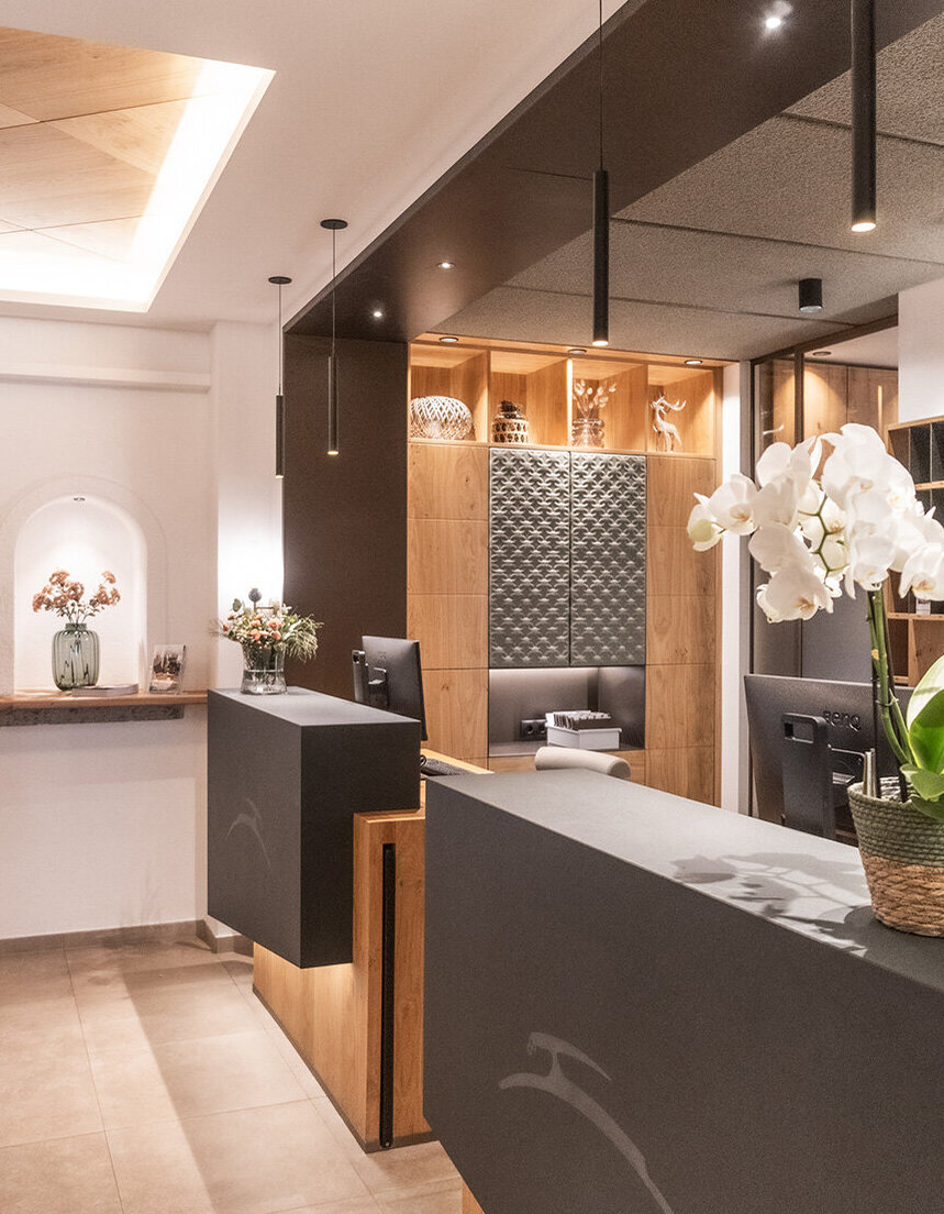 Moderner Empfangsbereich mit zwei schwarzen Schreibtischen, Holzakzenten, einem Korb mit weißen Orchideen, Pendelleuchten und dekorativen Regalen im Hintergrund. An der linken Wand befindet sich eine kleine Theke mit Blumen.