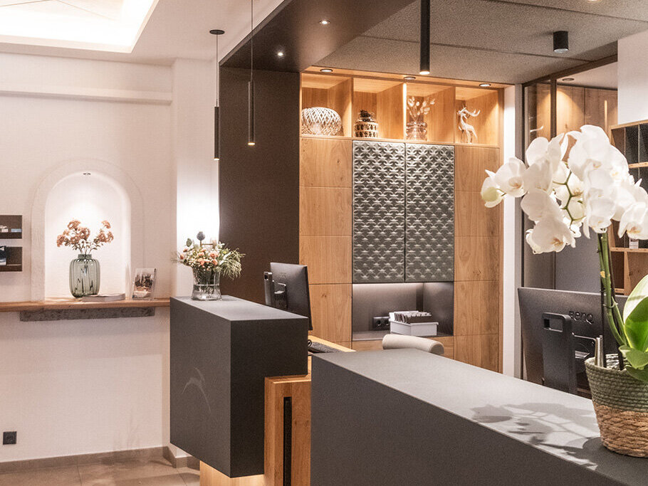 Moderner Empfangsbereich mit zwei schwarzen Schreibtischen, Holzakzenten, einem Korb mit weißen Orchideen, Pendelleuchten und dekorativen Regalen im Hintergrund. An der linken Wand befindet sich eine kleine Theke mit Blumen.