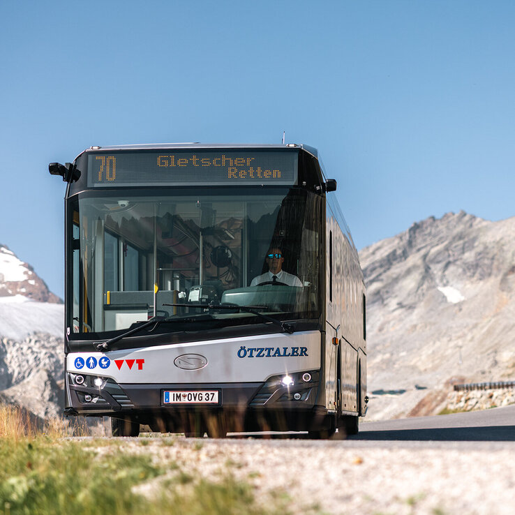 Ein weißer Linienbus mit der Linie 70 „Gletscher Retten“ fährt unter strahlend blauem Himmel eine Bergstraße entlang, im Hintergrund ragen felsige Gipfel empor.