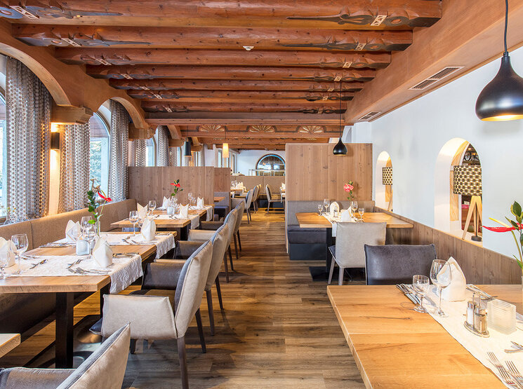 Ein helles, modernes Restaurant mit Holzbalken, hellen Holzböden, gedeckten Tischen mit weißen Servietten, Blumen in Vasen, gepolsterten Stühlen, Pendelleuchten und großen Bogenfenstern, durch die Tageslicht hereinströmt.