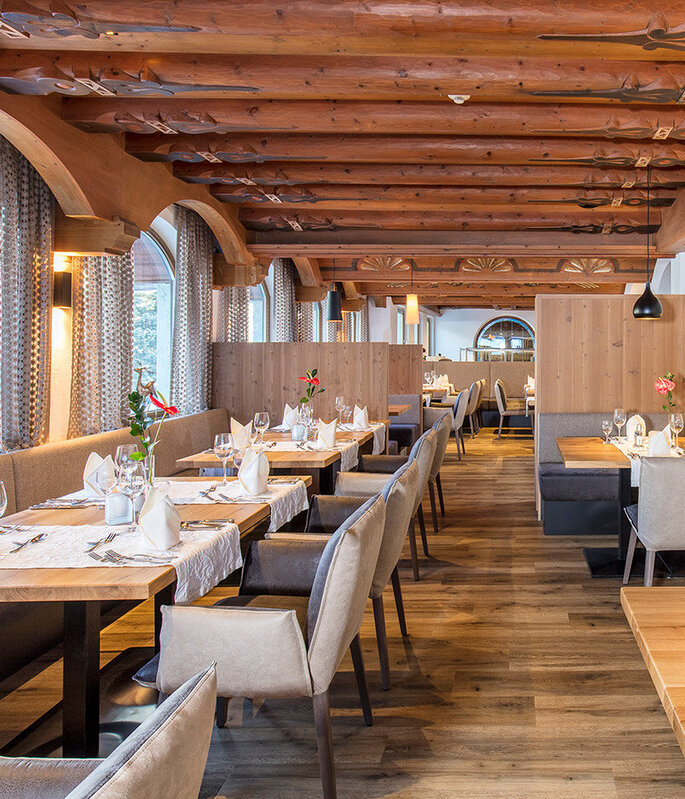 Ein helles, modernes Restaurant mit Holzbalken, hellen Holzböden, gedeckten Tischen mit weißen Servietten, Blumen in Vasen, gepolsterten Stühlen, Pendelleuchten und großen Bogenfenstern, durch die Tageslicht hereinströmt.