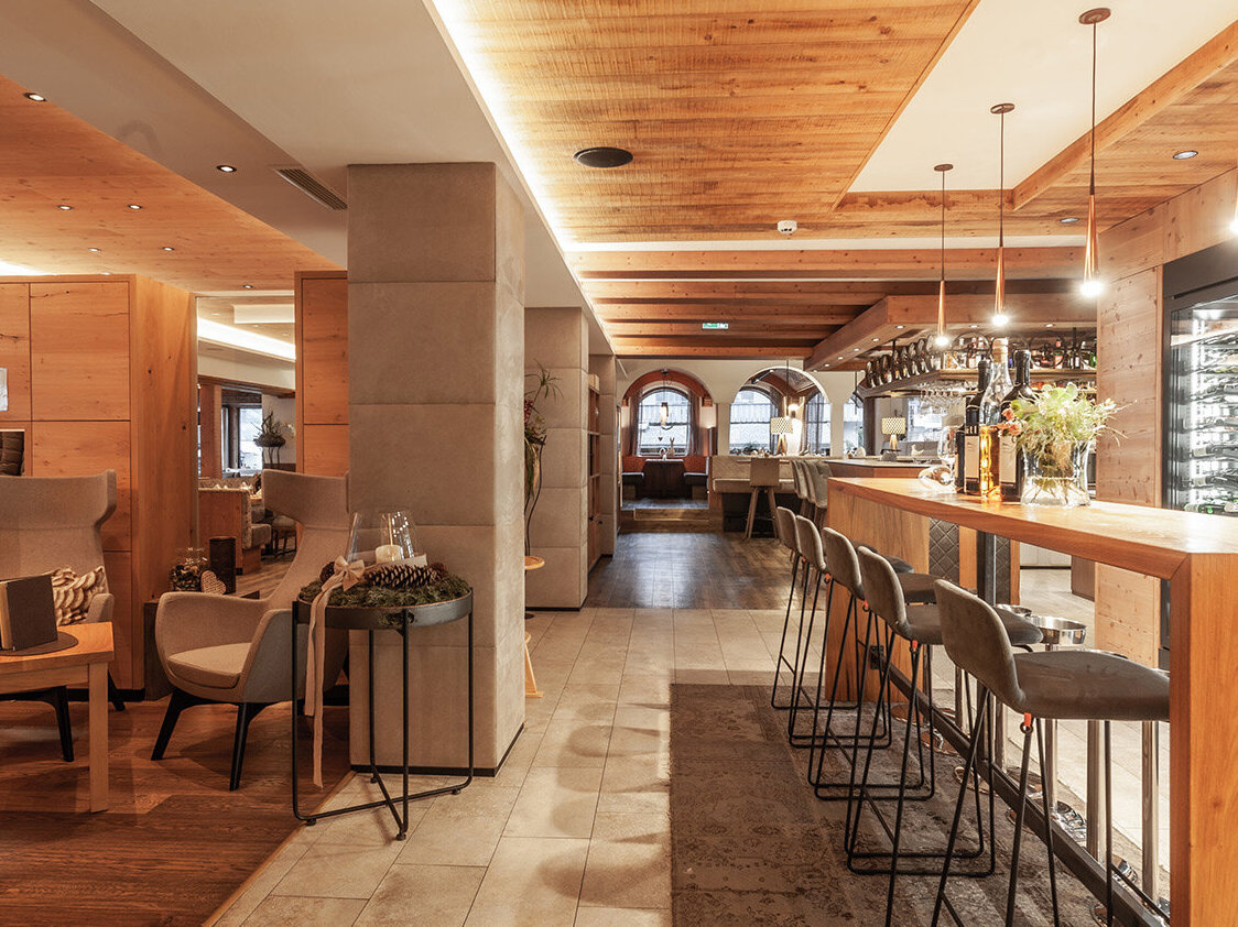 Modernes Restaurantinterieur mit Holzwänden und -decke, hohen Barhockern, Sesseln und warmer Beleuchtung. In einer Glasvitrine sind Weinflaschen ausgestellt, was eine gemütliche und einladende Atmosphäre schafft.