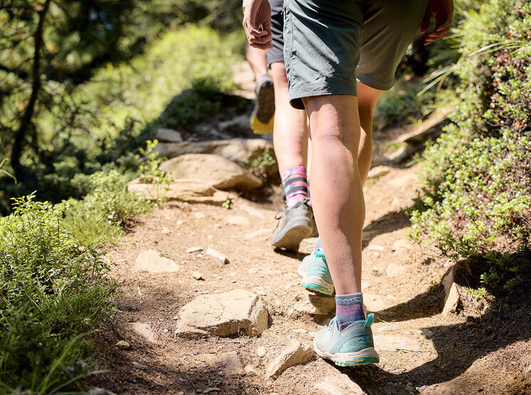 Zwei Menschen wandern auf einem felsigen, sonnenbeschienenen Weg, der von Grün umgeben ist. Man sieht nur ihre Beine und Füße, die mit Shorts, Socken und Sportschuhen bekleidet sind. Der Weg ist uneben, mit Felsen und Sonnenflecken.