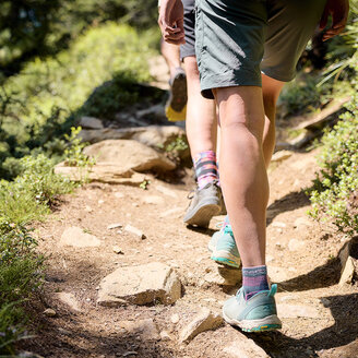 Zwei Menschen wandern auf einem felsigen, sonnenbeschienenen Weg, der von Grün umgeben ist. Man sieht nur ihre Beine und Füße, die mit Shorts, Socken und Sportschuhen bekleidet sind. Der Weg ist uneben, mit Felsen und Sonnenflecken.