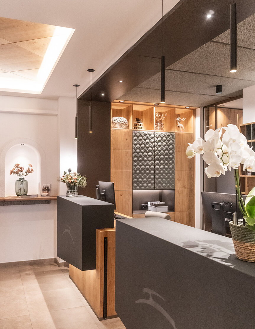 Ein moderner Empfangsbereich mit zwei schwarzen Theken, Holzakzenten, Orchideen in Töpfen, Pendelleuchten, einer Vase mit Blumen und sanfter Stimmungsbeleuchtung, die eine einladende Atmosphäre schafft.