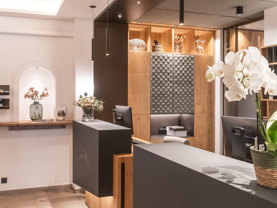 Ein moderner Empfangsbereich mit zwei schwarzen Theken, Holzakzenten, Orchideen in Töpfen, Pendelleuchten, einer Vase mit Blumen und sanfter Stimmungsbeleuchtung, die eine einladende Atmosphäre schafft.