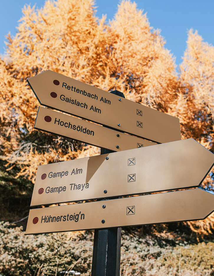Ein Wegweiser mit fünf gelben Richtungsschildern weist auf verschiedene Wanderwege hin, vor dem Hintergrund herbstlicher Bäume mit gold-orangefarbenem Laub unter einem strahlend blauen Himmel.