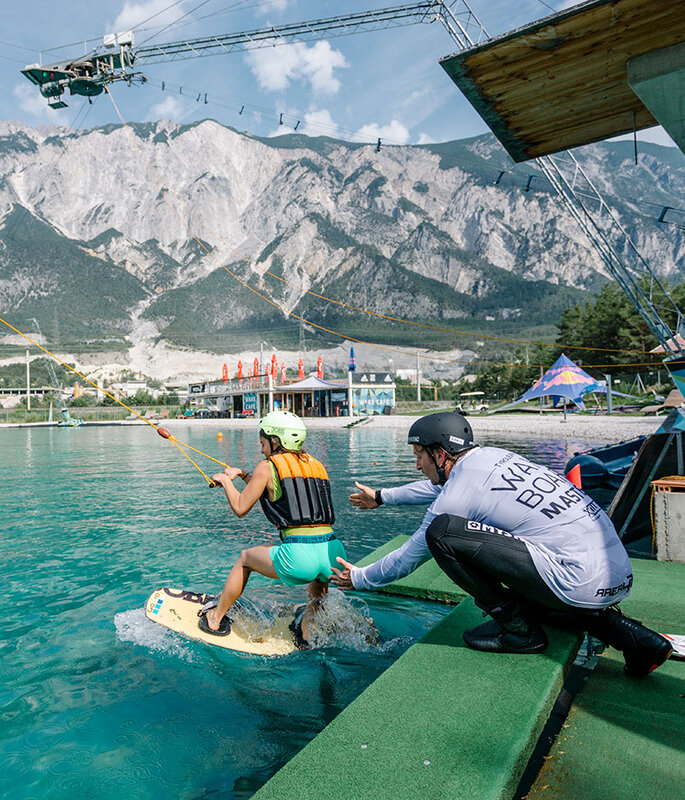 Eine Person auf einem Wakeboard wird an einem Seil über türkisfarbenes Wasser gezogen, im Hintergrund sind Berge zu sehen. Ein Lehrer kniet in der Nähe, während andere von einem Steg aus zuschauen.