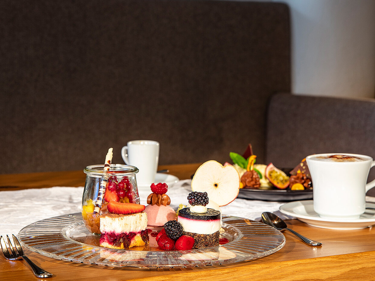 Eine Glasplatte mit verschiedenen Desserts und Beeren auf einem Holztisch, daneben eine Tasse Kaffee, eine Untertasse, ein Löffel und ein kleines Dessertglas, im Hintergrund ist ein weiterer Dessertteller zu sehen.