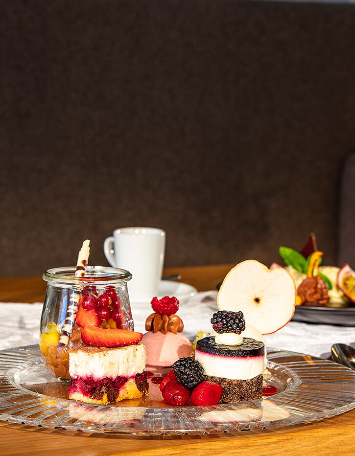 Eine Glasplatte mit verschiedenen Desserts und Beeren auf einem Holztisch, daneben eine Tasse Kaffee, eine Untertasse, ein Löffel und ein kleines Dessertglas, im Hintergrund ist ein weiterer Dessertteller zu sehen.
