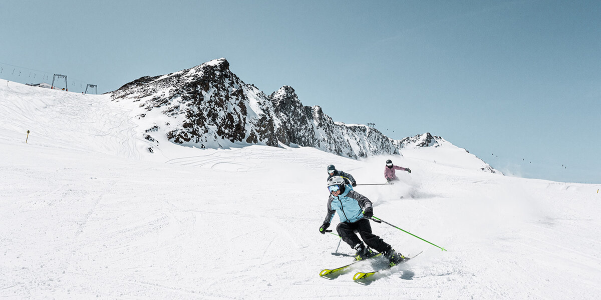 Drei Personen fahren unter strahlend blauem Himmel auf einem verschneiten Berghang Ski, im Hintergrund sind felsige Gipfel zu sehen.
