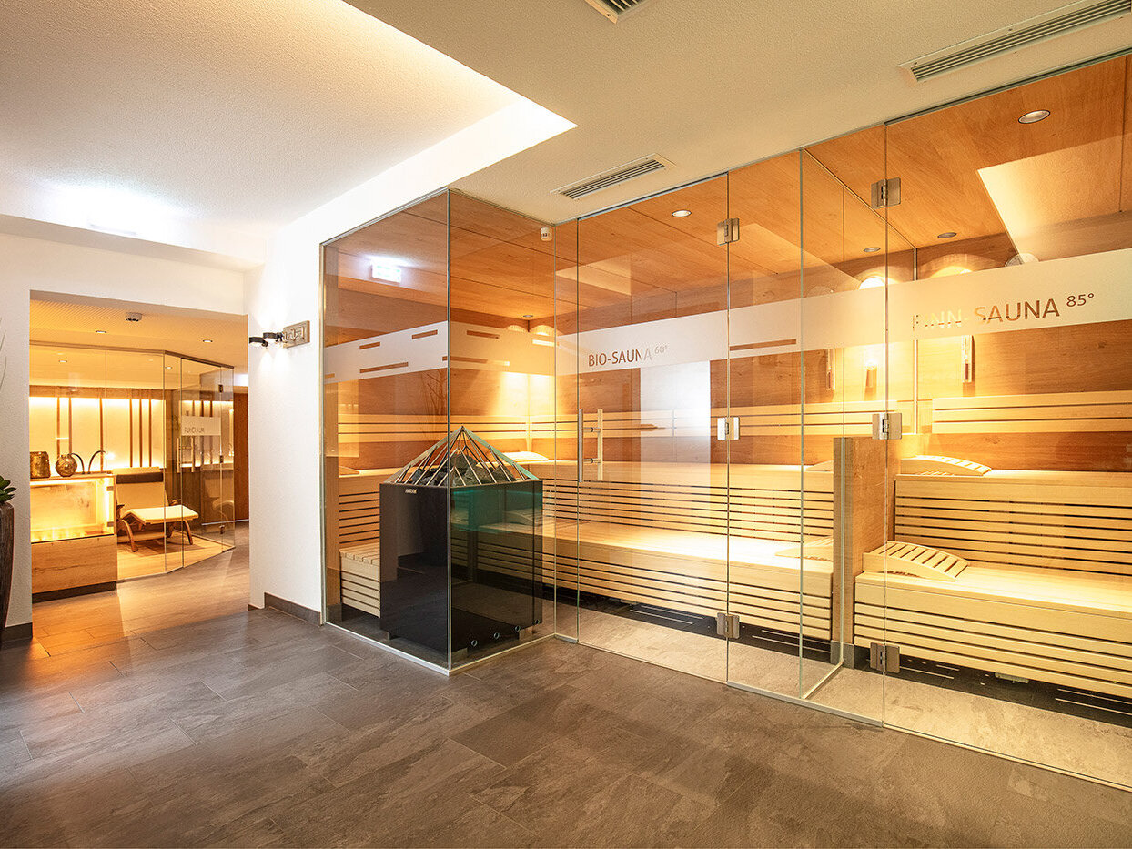 Ein moderner Wellnessbereich mit einer Sauna mit Glaswänden auf der rechten Seite, Holzbänken im Inneren und einer Entspannungslounge mit Sesseln, die durch eine offene Tür auf der linken Seite zu sehen sind.