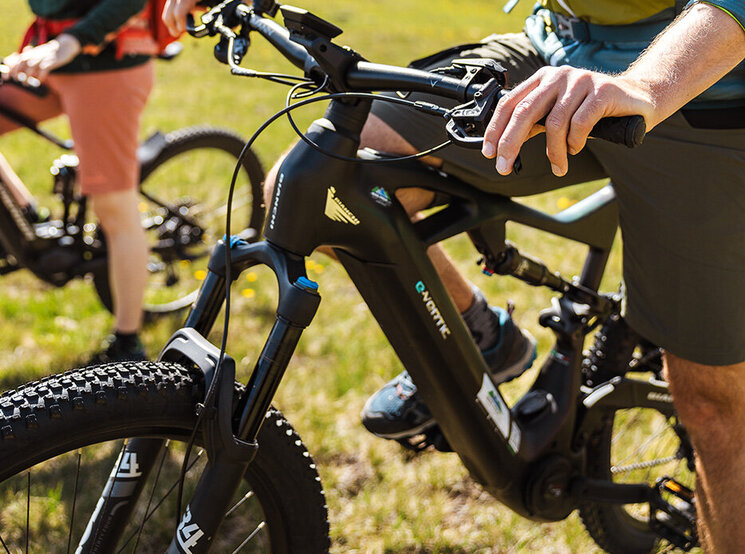 Zwei Personen mit Helmen und Outdoor-Ausrüstung fahren mit Elektro-Mountainbikes auf einem grasbewachsenen Weg, im Hintergrund sind Berge zu sehen. Eine der beiden lächelt die andere an und genießt den sonnigen Tag im Freien.
