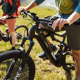 Zwei Personen mit Helmen und Outdoor-Ausrüstung fahren mit Elektro-Mountainbikes auf einem grasbewachsenen Weg, im Hintergrund sind Berge zu sehen. Eine der beiden lächelt die andere an und genießt den sonnigen Tag im Freien.