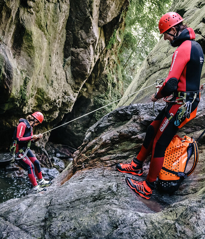Zwei Personen in roten Neoprenanzügen und Helmen betreiben Canyoning. Eine seilt sich an einer Felswand in der Nähe des Wassers ab, während die andere das Seil sichert und dabei von Felsen und üppiger Vegetation umgeben ist.