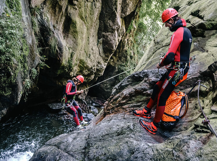 Zwei Personen in roten Neoprenanzügen und Helmen betreiben Canyoning. Eine seilt sich an einer Felswand in der Nähe des Wassers ab, während die andere das Seil sichert und dabei von Felsen und üppiger Vegetation umgeben ist.