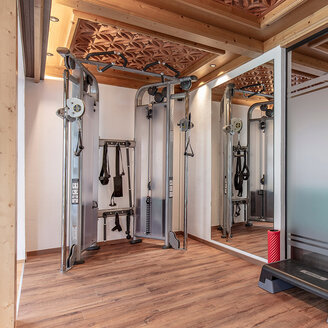 Ein kleiner Fitnessraum mit Holzboden, einer Doppel-Kabelmaschine, einem Wandspiegel, einer Step-Plattform und Milchglasplatten. Die Decke ist mit dekorativen Holzschnitzereien verziert.