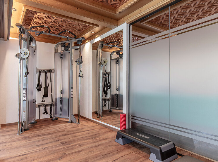 Ein kleiner Fitnessraum mit Holzboden, einer Doppel-Kabelmaschine, einem Wandspiegel, einer Step-Plattform und Milchglasplatten. Die Decke ist mit dekorativen Holzschnitzereien verziert.