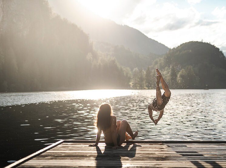 Zwei Menschen auf einem Steg am See: Der eine sitzt mit den Beinen im Wasser, während der andere vor dem Hintergrund von Bergen und Sonnenschein in die Luft springt und in den See taucht.