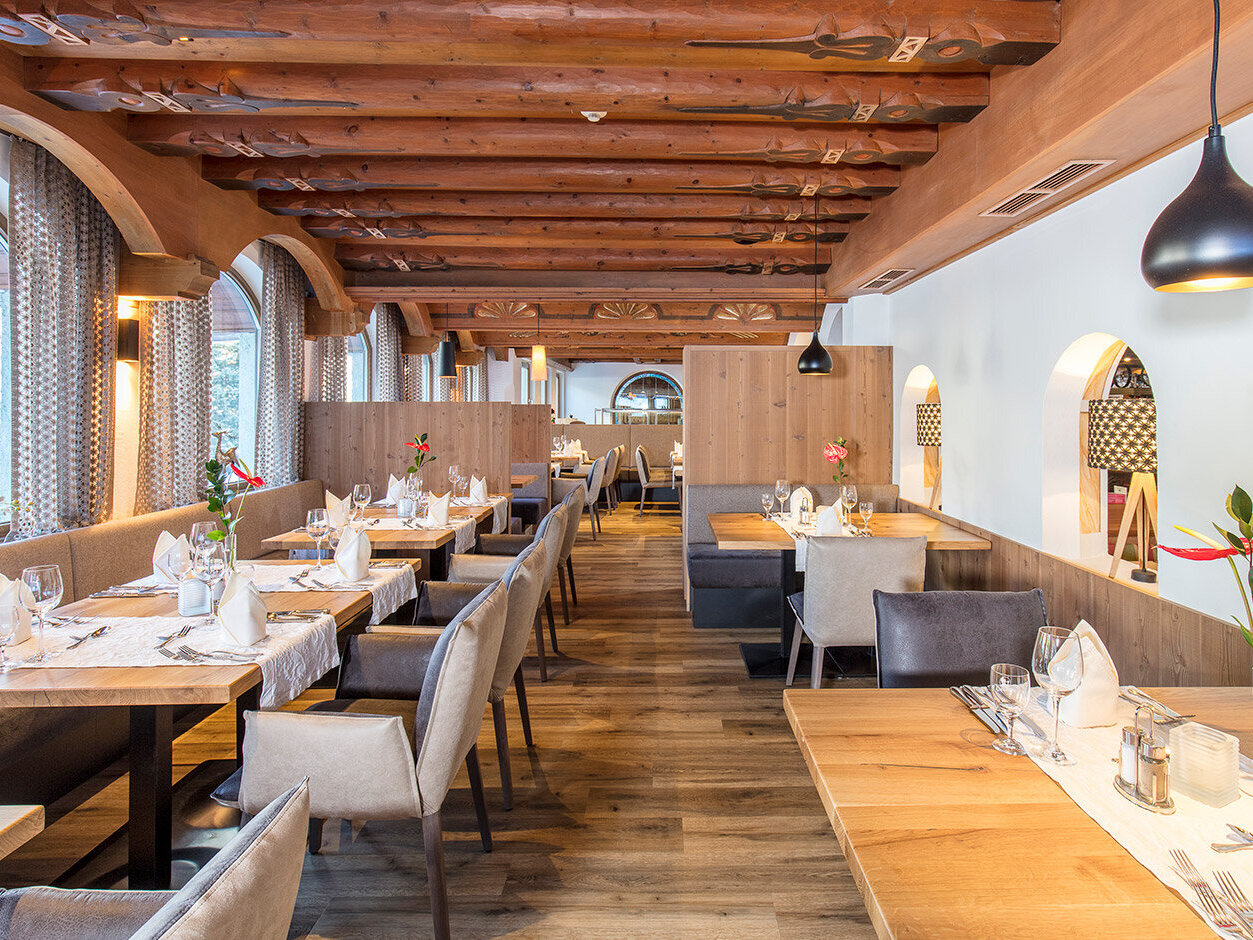 Ein modernes Restaurant mit Holzdecken und -böden, gepflegt gedeckten Tischen mit weißen Servietten und Blumen, gepolsterten Stühlen, sanfter Beleuchtung und großen Bogenfenstern, durch die Tageslicht hereinströmt.