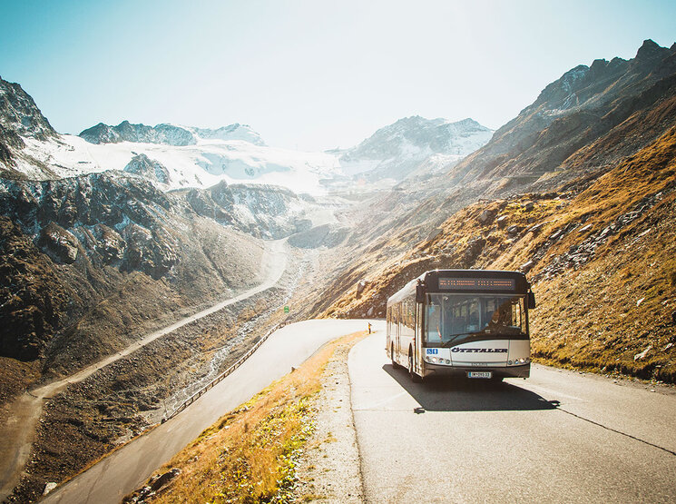 Ein Bus fährt auf einer kurvenreichen Bergstraße, umgeben von schroffen, schneebedeckten Gipfeln und einem klaren Himmel.