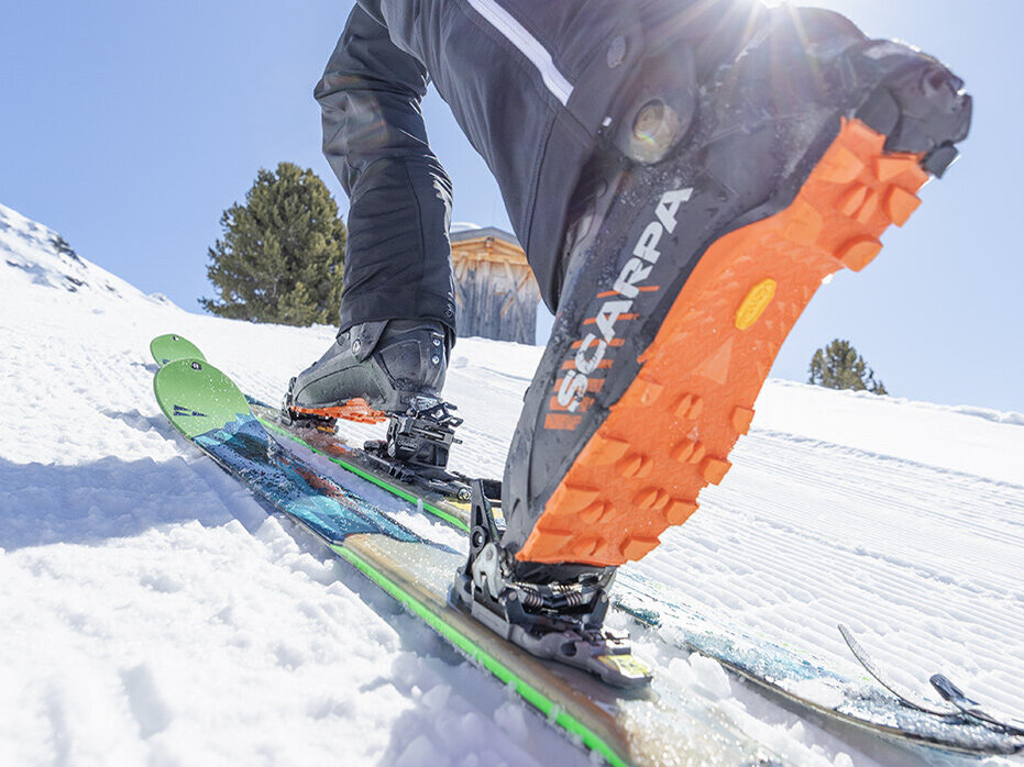 Nahaufnahme eines Skischuhs und von Skiern, im Hintergrund scheint helles Sonnenlicht. Der Skifahrer bewegt sich auf verschneitem Gelände bergauf; er trägt eine schwarze Hose. Im Bild sind Kiefern und ein blauer Himmel zu sehen.