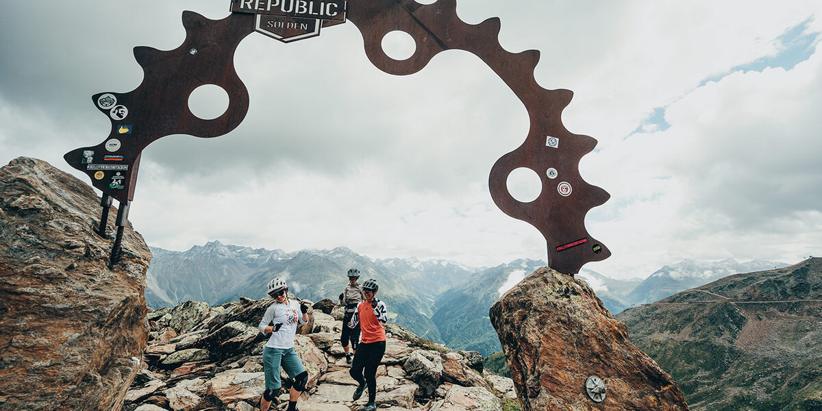 Drei Radfahrer stehen auf einem felsigen Berggipfel unter einem großen Metallschild mit der Aufschrift „Bike Republic Sölden“, das die Form eines Fahrradkettenrads hat, im Hintergrund sind Berge und ein bewölkter Himmel zu sehen.