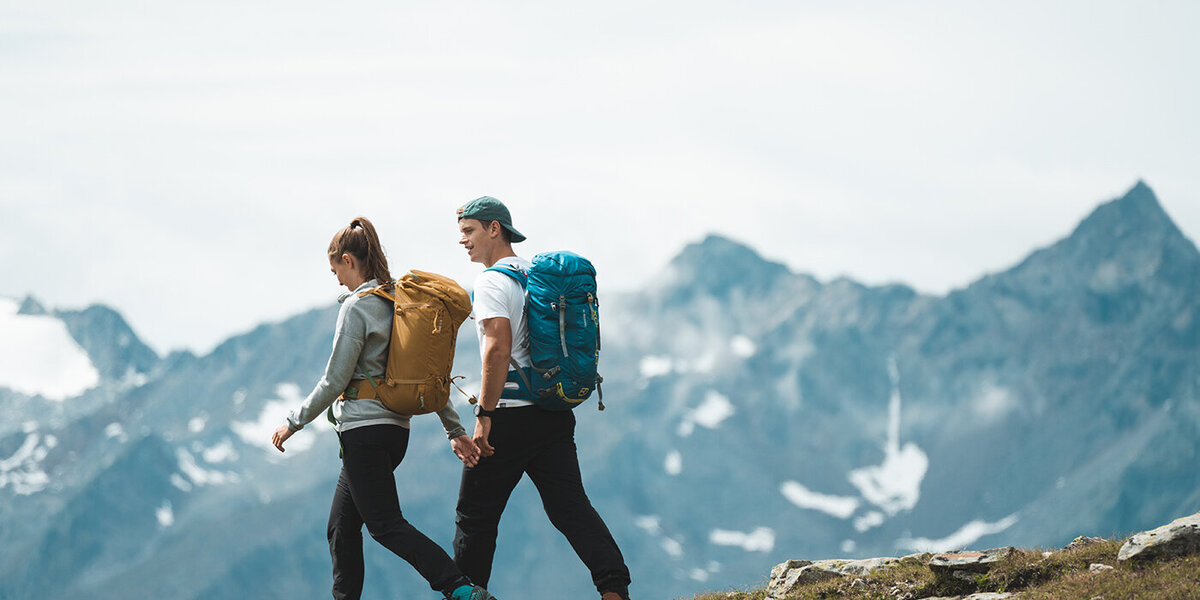 Zwei Menschen mit Rucksäcken wandern im Freien auf einem grasbewachsenen Hang, im Hintergrund sind schneebedeckte, schroffe Berge zu sehen, über ihnen ein bewölkter Himmel.