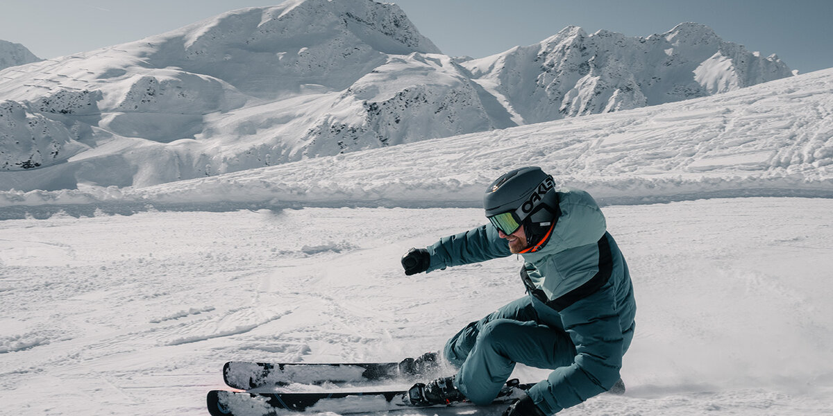 Ein Skifahrer in blauer Ausrüstung und mit Helm fährt unter klarem Himmel eine scharfe Kurve auf einem verschneiten Berghang, im Hintergrund sind schneebedeckte Gipfel zu sehen.