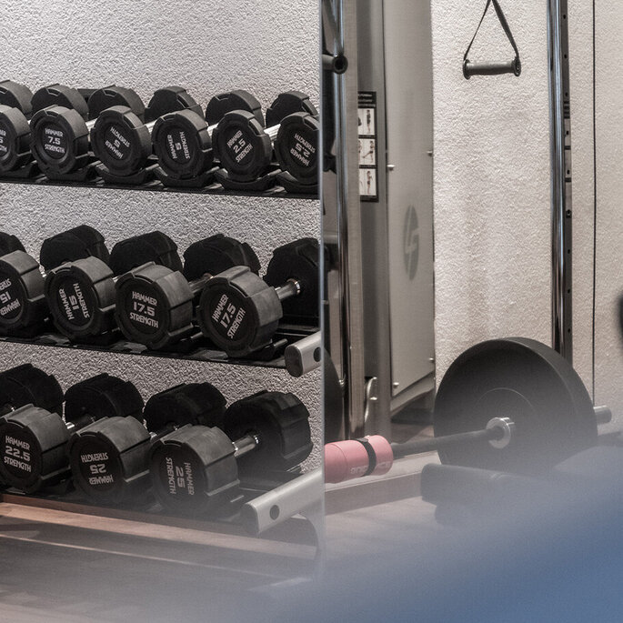 Eine Szene in einem Fitnessstudio mit einem Hantelständer, einer Kabelzugmaschine, einem Griffaufsatz und einer Langhantel mit Gewichten. Der Raum wirkt sauber und aufgeräumt, wobei einige Fitnessgeräte teilweise zu sehen sind.