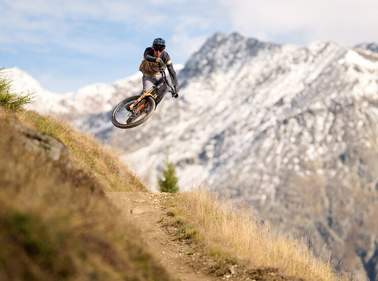 Ein Mountainbiker springt von einem Feldweg ab, lehnt sich dabei in der Luft zur Seite, im Hintergrund sind schneebedeckte Berge und ein blauer Himmel zu sehen.