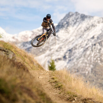 Ein Mountainbiker springt von einem Feldweg ab, lehnt sich dabei in der Luft zur Seite, im Hintergrund sind schneebedeckte Berge und ein blauer Himmel zu sehen.