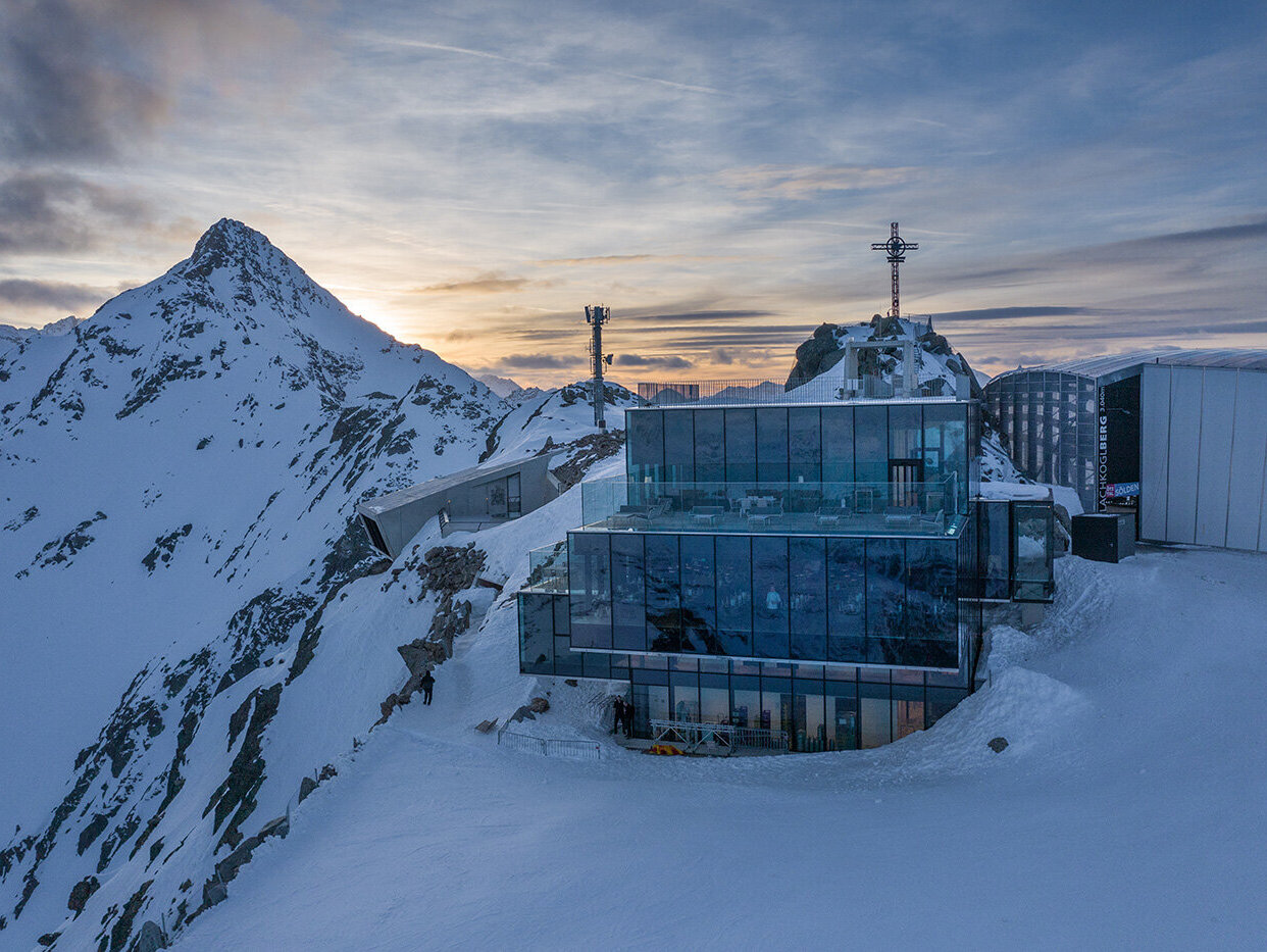 Ein modernes Glasgebäude, das iceQ, thront bei Sonnenuntergang auf einem schneebedeckten Berggipfel, umgeben von verschneiten Hängen, mit Berggipfeln und einem teilweise bewölkten Himmel im Hintergrund.
