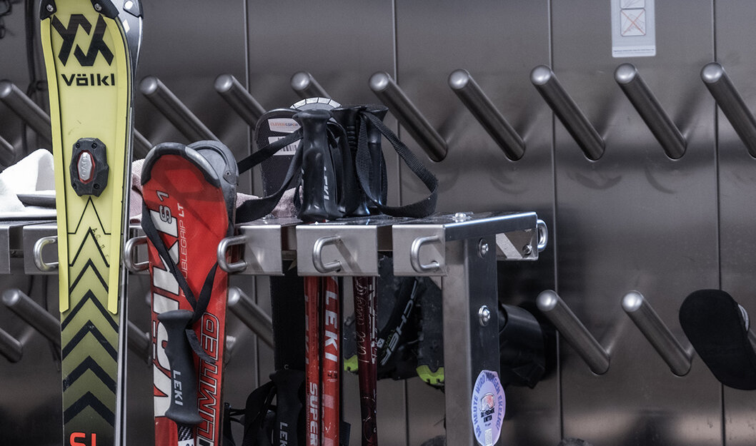 Eine Nahaufnahme eines Skiraums, in dem Skier, Skistöcke, Skischuhe und schwarze Handschuhe an einem Metallgestell und an Wandhaltern hängen. Der Raum wirkt aufgeräumt und bereit für den Wintersport.