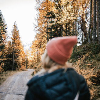 Eine Person mit einer rosa Mütze und einer dunklen Jacke geht einen Feldweg durch einen Wald mit herbstlich gefärbten Bäumen entlang und blickt dabei nach vorne. Die Szene wirkt hell und friedlich.