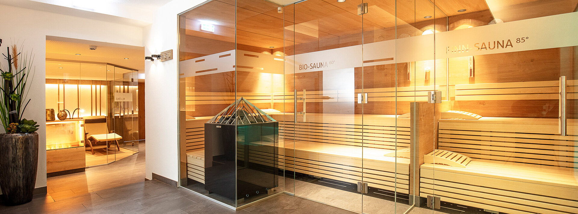 Moderner Wellnessbereich mit glaswandigen Saunen, die mit Holzbänken, einem Ofen und warmer Beleuchtung ausgestattet sind. Durch eine offene Tür im Hintergrund ist ein Loungebereich mit Stühlen und Pflanzen zu sehen.
