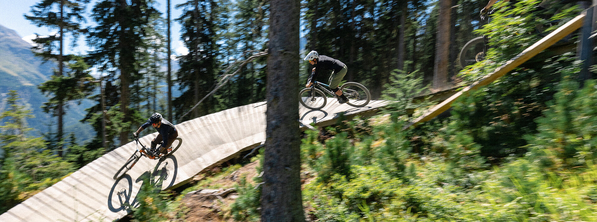 Zwei Radfahrer mit Helmen fahren einen kurvenreichen Holzpfad in einer bewaldeten Berglandschaft hinunter, umgeben von Bäumen und Grün, während das Sonnenlicht durch die Äste scheint.
