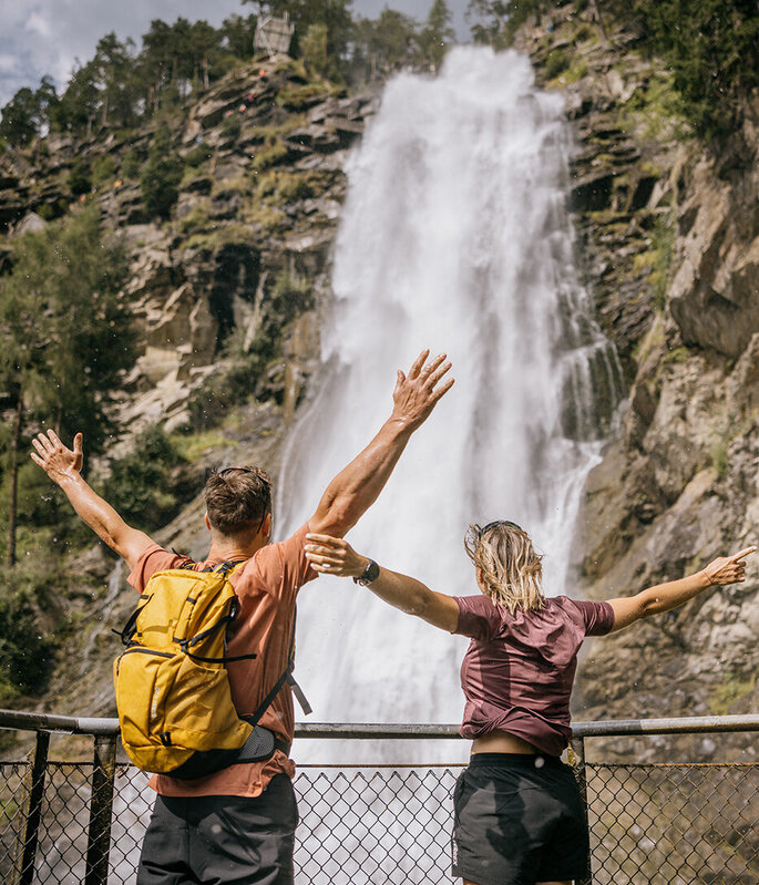 Zwei Personen stehen mit erhobenen Armen vor einem großen, rauschenden Wasserfall, von hinten gesehen. Eine der beiden trägt einen gelben Rucksack. Beide genießen die malerische Aussicht, umgeben von Bäumen und felsigen Klippen.