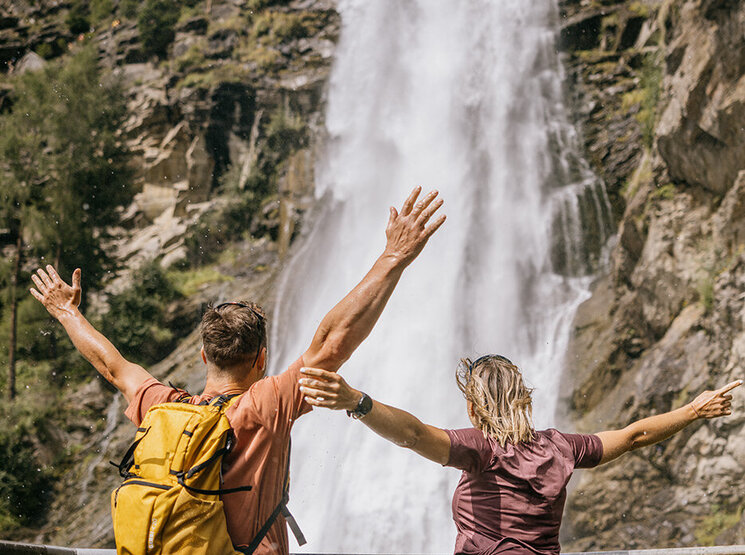 Zwei Personen stehen mit erhobenen Armen vor einem großen, rauschenden Wasserfall, von hinten gesehen. Eine der beiden trägt einen gelben Rucksack. Beide genießen die malerische Aussicht, umgeben von Bäumen und felsigen Klippen.