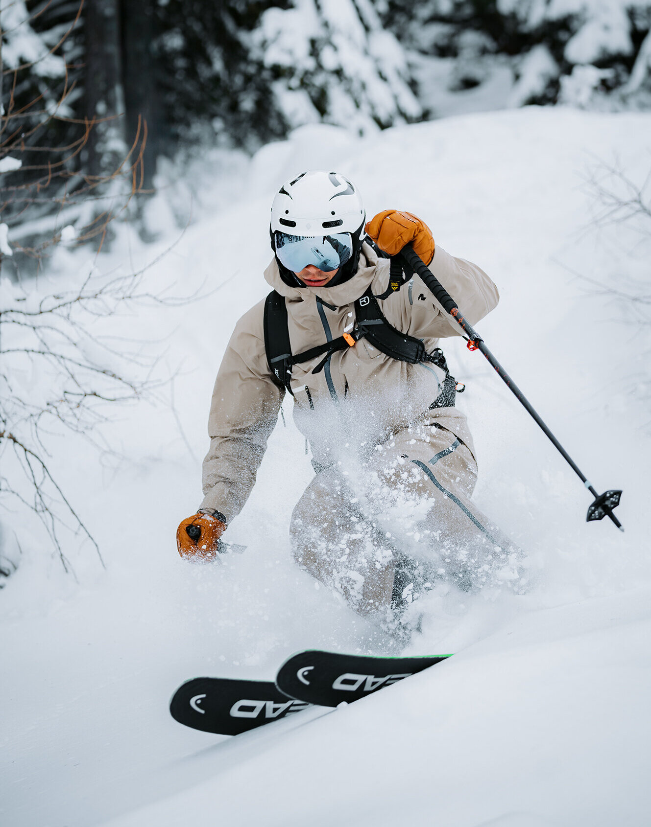 Ein Skifahrer in beiger Skikleidung und mit Helm fährt auf einem verschneiten, von Bäumen gesäumten Hang durch frischen Pulverschnee; er hält Skistöcke in den Händen und trägt eine Skibrille.