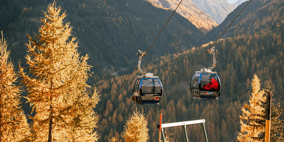 Zwei Gondelbahnen gleiten über einen Wald mit gelb gefärbten Herbstbäumen hinweg, im Hintergrund ragen hohe Berge empor, und Sonnenlicht taucht die Landschaft in ein warmes Licht.