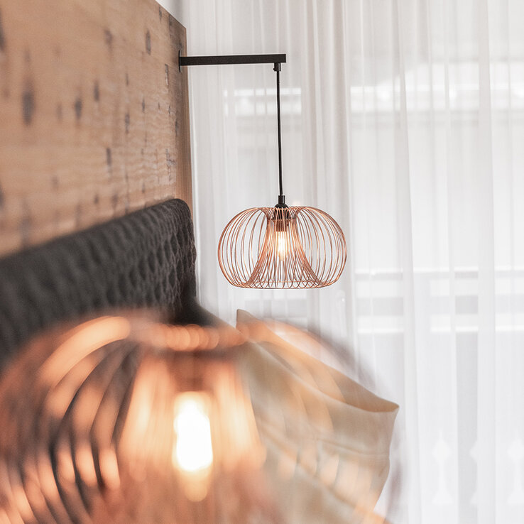 Nahaufnahme von zwei modernen Pendelleuchten mit Drahtgestell, die neben einem Bett mit gepolstertem Kopfteil hängen. Durchsichtige weiße Vorhänge verdecken ein Fenster in dem sanft beleuchteten, modernen Schlafzimmer.