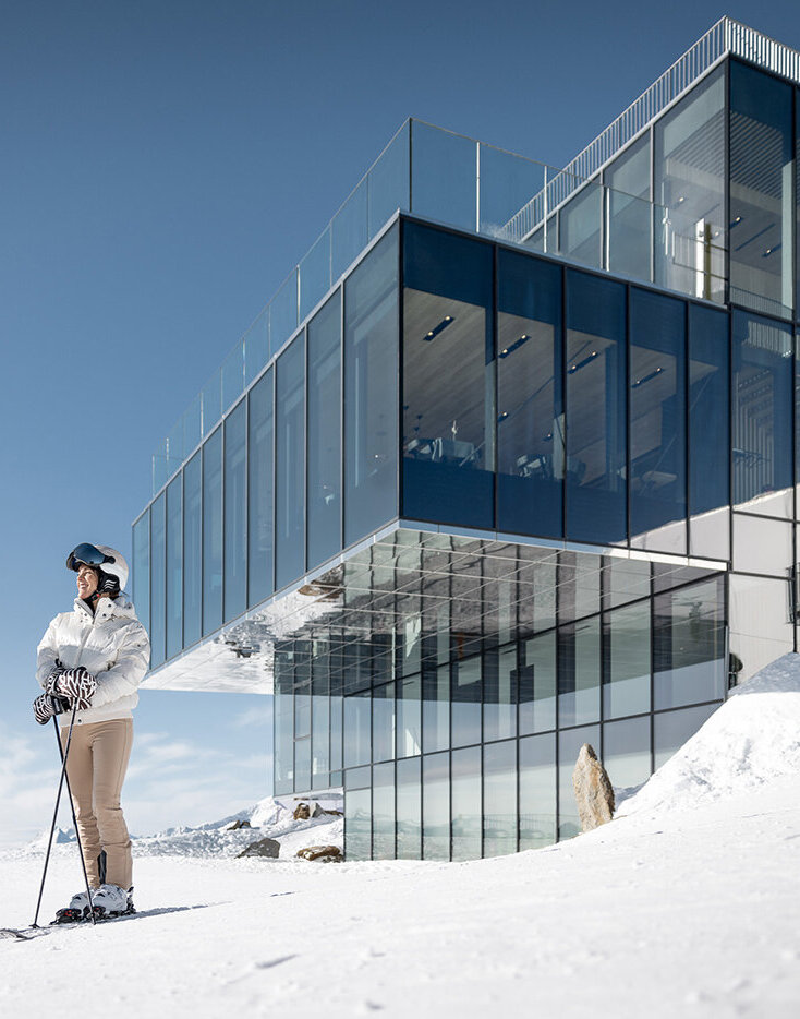 Zwei Personen in Skikleidung stehen im Schnee in der Nähe eines modernen Glasgebäudes, im Hintergrund sind unter einem strahlend blauen Himmel Berge zu sehen.