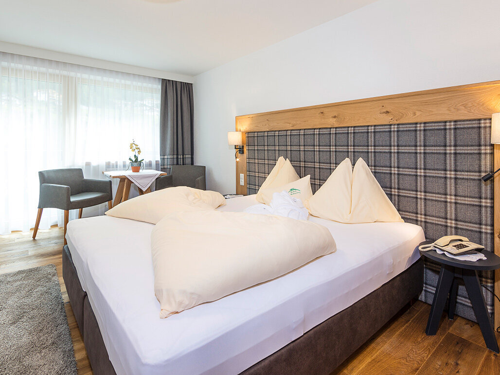 Ein helles Hotelzimmer mit einem großen Bett, einem karierten Kopfteil, zwei beigen Stühlen an einem runden Tisch am Fenster, einem Holzboden, einem grauen Teppich und einem Nachttisch mit einem Telefon und einer Lampe.