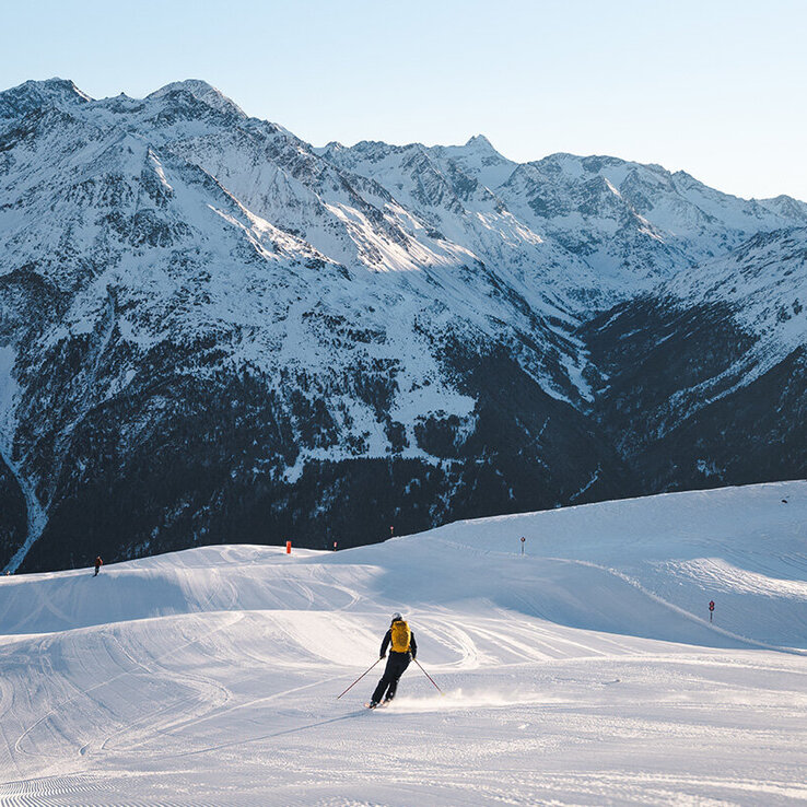 Ein Skifahrer in einer gelben Jacke fährt einen präparierten, verschneiten Hang hinunter, umgeben von hohen, schroffen Bergen unter einem klaren Himmel.