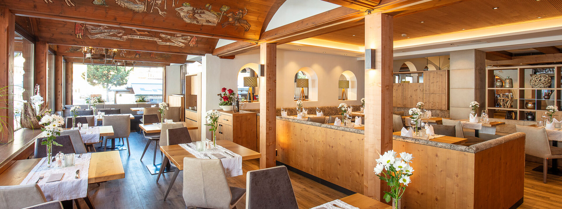 Ein helles, modernes Restaurant mit Holzmöbeln, mit Blumen geschmückten Tischen, gepolsterten Stühlen und natürlichem Licht, das durch große Fenster hereinströmt. An der Holzdecke sind kunstvolle Verzierungen zu sehen.